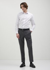 Mann steht mit den Händen in den Taschen, trägt ein weißes Hemd, graue karierte Hose, einen schwarzen Gürtel und schwarze Slipper vor einem einfarbigen Hintergrund.