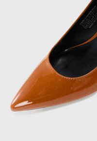 Chaussure à talon haut en cuir verni marron avec un bout pointu et un intérieur noir. Présente une finition lisse et brillante ainsi que des détails de couture subtils.