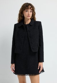 Sort tweedjakke med krave, frontknapper og en struktureret pasform, parret med en matchende kjole med frynsedetaljer i kanten.