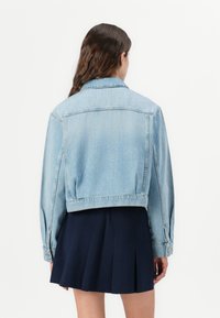 Lys blå denimjakke med en avkortet design, synlige sømmer og subtile dekorasjoner. Stoffet har en klassisk, myk tekstur.
