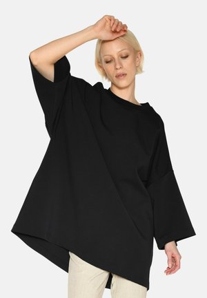 Čierne oversize tričko z mäkkého materiálu s širokým výstrihom, spadnutými ramenami a dlhším zadným lemom, nosené cez svetlé nohavice.