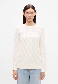 Pletený sveter v slonovinovej farbe s textúrovaným káblovým vzorom, okrúhlym výstrihom a rebrovanými manžetami a spodným lemom. Nosený s svetlomodrými džínami.