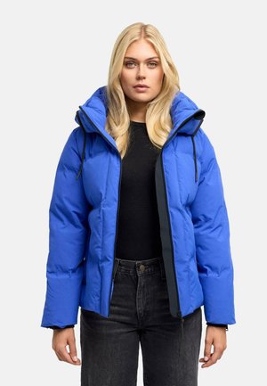Femme blonde portant une doudoune bleue par-dessus un t-shirt noir et un jean foncé, regardant droit devant avec une expression neutre.