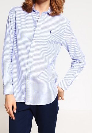 Personne portant une chemise boutonnée rayée verticalement bleu et blanc à manches longues et un pantalon bleu foncé.