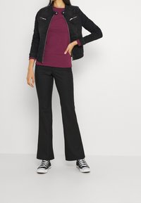 Schwarze Jeansjacke mit Reißverschlusstaschen, lila gerippter Oberteil und schwarze ausgestellte Hosen. Schwarz-weiße Sneakers runden das Outfit ab.