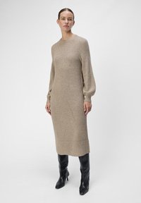 Beige gebreide jurk met lange ballonmouwen, geribbelde textuur en een rechte silhouet. Gecombineerd met zwarte laarzen tot de knie.