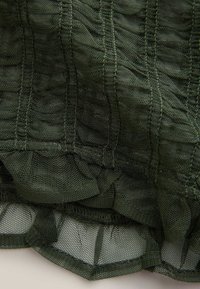 Przezroczysta, oliwkowozielona tkanina z teksturowanymi falbankami. Materiał ma siateczkową strukturę, widoczne przeszycia i warstwowe krawędzie.