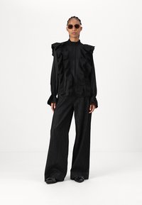 Blusa negra con detalles de volantes, mangas abullonadas y cuello alto, combinada con pantalones de mezclilla negros de pierna ancha y zapatos planos negros.