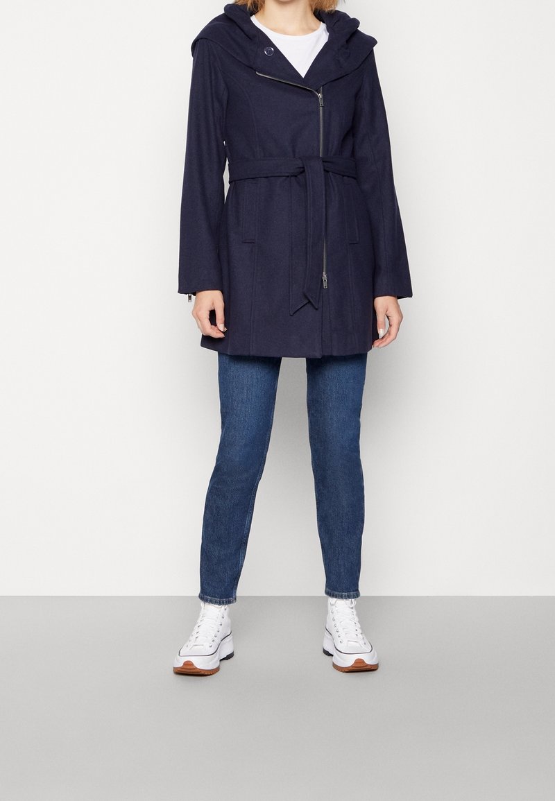 Cappotto blu navy con zip, ampio cappuccio, cintura in vita e due tasche frontali; indossato sopra una maglietta bianca e jeans blu.