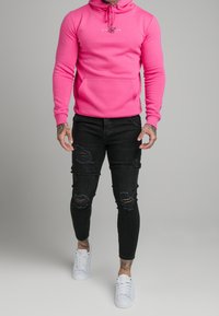 Ljusrosa hoodie med en frontficka och dragsko, kombinerad med slitna svarta skinny jeans och vita sneakers.