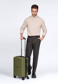 Hombre con un suéter de cuello alto beige y pantalones oscuros, de pie con una mano en el bolsillo y la otra sosteniendo el asa de una maleta verde oliva.