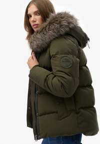 Chaqueta acolchada verde oliva con capucha de piel sintética, cierre de cremallera y tejido texturizado. Presenta un logo circular en la manga izquierda.