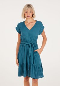 Teal jurk met korte mouwen, V-hals, bevestigde taille en strikband; heeft een gelaagde rok en een subtiele textuur.