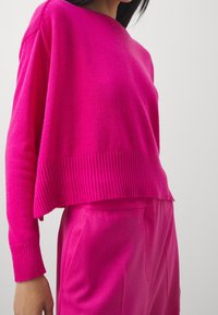 Fuchsia gestrickter Pullover mit einem gerippten Saum, weiten Ärmeln und einer kastigen Passform, kombiniert mit passenden, maßgeschneiderten Hosen. Glatte Textur überall.