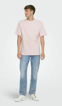 Light pink cotton t-shirt with short sleeves, crew neck, paired with light blue denim jeans and white sneakers. Model stands sideways.