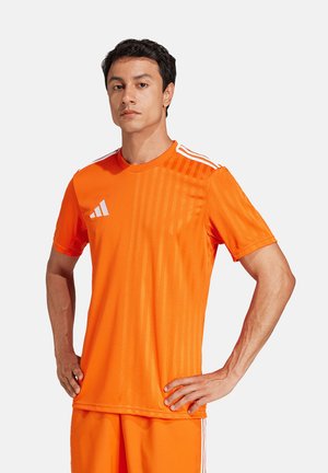 Mann steht mit den Händen in den Hüften, trägt leuchtend orange Adidas-Sportbekleidung mit drei weißen Streifen auf den Schultern und Logo auf der Brust.