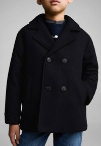 Svart ull peacoat med dubbelknäppt design, slagkrage, två fickor och rund svart knappar, i kombination med en blå tröja och jeans.