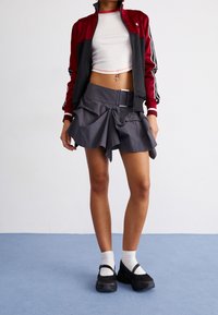 Rote und schwarze Jacke mit Reißverschluss, weißes cropped Top, gestuftem grauer Rock und schwarze Schuhe mit weißen Socken. Strukturierter Stoff und geschichtetes Design.