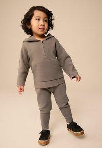 Grå hoodie och byxor i mjukt tyg med dragkedja framtill, som har en framficka. Svarta skor med ljusbruna sulor.