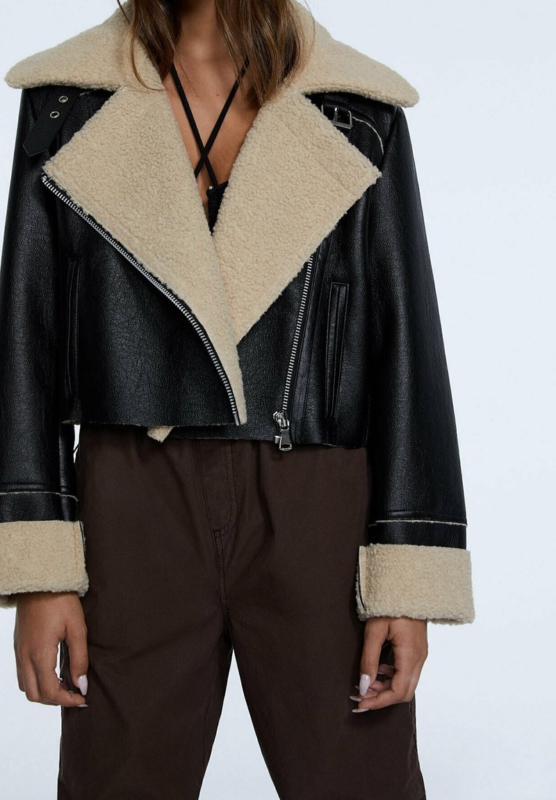 Veste noire courte avec un col et des manchettes en fausse fourrure beige. Présente des fermetures éclair en argent et un extérieur en cuir texturé.