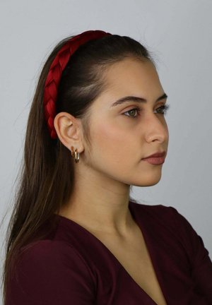 Czerwona pleciona opaska z tkaniny na ciemnobrązowych włosach, w towarzystwie złotych kolczyków kołowych, na neutralnym tle.