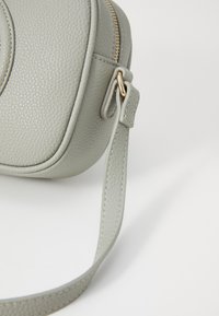 Γκρίζα δερμάτινη τσάντα χιαστί με τραχιά υφή, στρογγυλή μορφή, χρυσό φερμουάρ και αποσπώμενο λουρί. Διακοσμημένη με καθα�ρά ραψίματα.