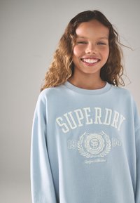 Šviesiai mėlynas džemperis su ilgomis rankovėmis, apvalia iškirpte, priekyje baltais rašmenimis užrašytas logotipas „SUPERDRY“, pasižymintis tekstūruota medžiaga ir siuvinėtomis detalėmis.