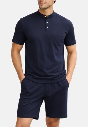 Mann trägt navyblaues Henley-Shirt mit kurzen Ärmeln und Knöpfen und passende navyblaue Shorts, Hände in den Taschen, vor einfachem Hintergrund.