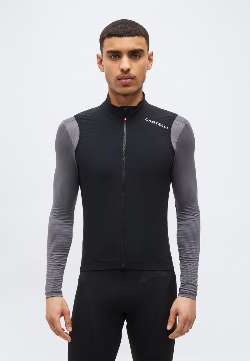 Άνδρας που φοράει μαύρο γιλέκο ποδηλασίας πάνω από γκρι μακρυμάνικη μπλούζα, στέκεται μπροστά σε απλό ανοιχτό γκρι φόντο.