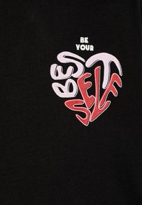 Svart stoff med et fargerikt "Vær deg selv" hjerte-design i rosa og rødt, med stilisert, uregelmessig skrift på forsiden.
