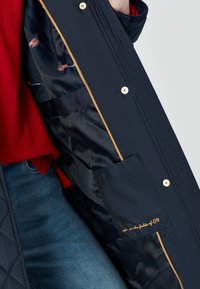 Una persona con un maglione rosso e jeans blu mostra l'interno di un cappotto blu navy con fodera a stampa di fagiano e una tasca interna con testo ricamato.