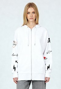 Felpa bianca con zip e dettagli grafici sulle maniche, tra cui cuori, un teschio e fulmini. Tessuto morbido; vestibilità comoda.