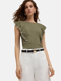 Olivgrön ribbad topp med volangärmar, vit kant, kombinerad med högmidjade vita byxor och ett svart bälte med ett guldspänne.