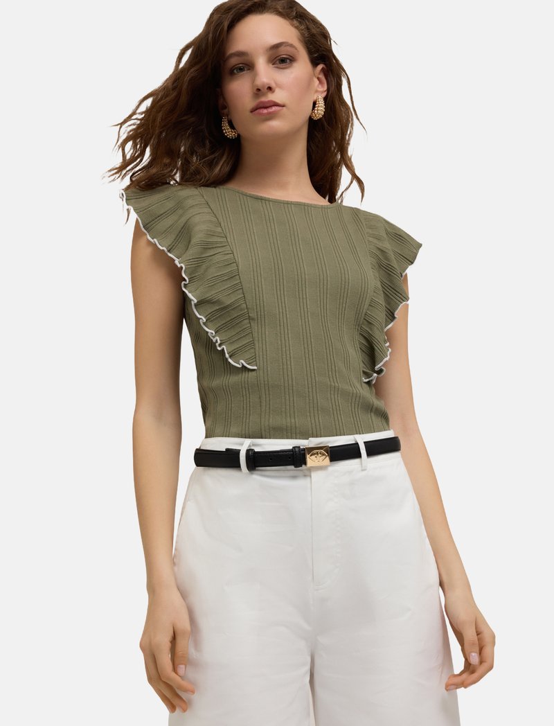 Olivgrön ribbad topp med volangärmar, vit kant, kombinerad med högmidjade vita byxor och ett svart bälte med ett guldspänne.
