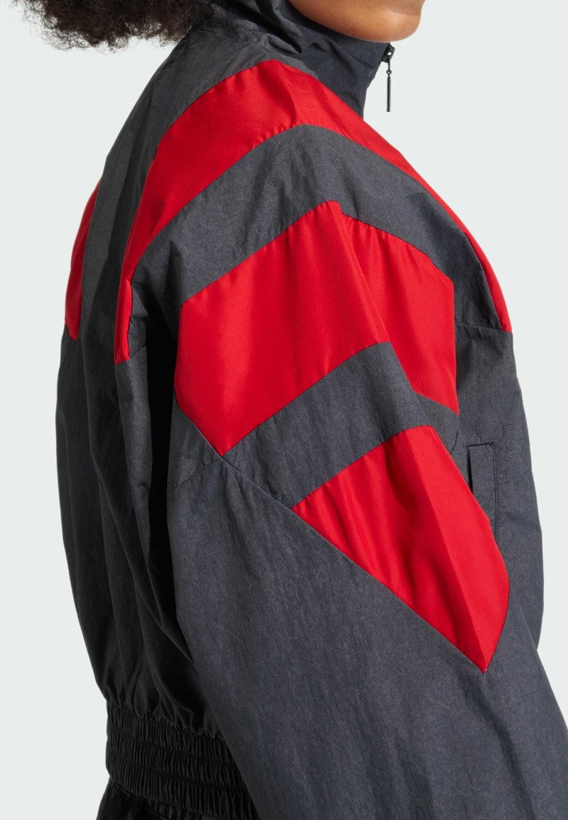 Schwarze und rote Windjacke mit hohem Kragen, elastischem Taillenbund und auffälligen, in Chevron-Optik gestalteten Farbblockstreifen an den Ärmeln.