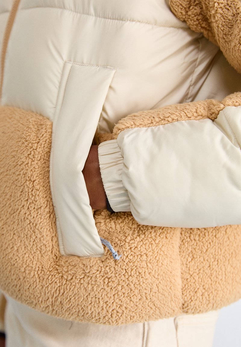 Kurtka puchowa w kolorach kremowym i karmelowym, z blokowym wzorem, charakteryzująca się miękkim, teksturowanym materiałem i elastycznymi mankietami. Widać boczną kieszeń.