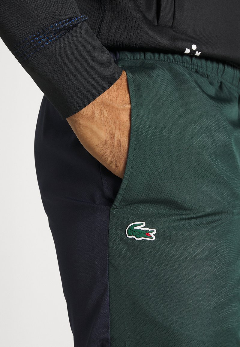 Pantalons de sport verts et bleu marine avec une finition texturée, dotés d'une poche latérale et d'un logo Lacoste blanc. Ceinture élastique visible.