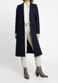 Cappotto lungo doppiopetto blu navy con bottoni neri, indossato sopra una camicetta color crema e pantaloni beige, abbinato a stivali alla caviglia in pelle di serpente.
