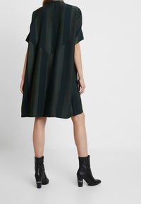 Abito in satin verde scuro con righe verticali, maniche corte e vestibilità ampia, abbinato a stivaletti neri con tacco alla caviglia caratterizzati da un tacco decorativo.