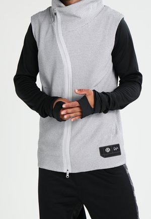 Osoba nosząca bezrękawnik o szarej, fakturowanej powierzchni z zamkiem z przodu na czarnej koszuli z długim rękawem, ręce złożone na wysokości talii.