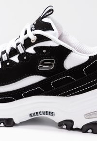 Black and white sneaker with suede and mesh materials. Features a prominent silver logo, white laces, and textured rubber sole with black accents.