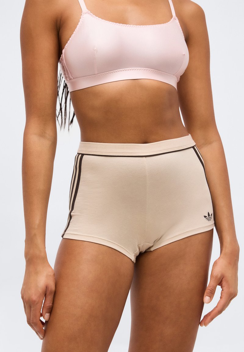Γυναίκα που φοράει ανοιχτό ροζ αθλητικό σουτιέν και μπεζ σορτς Adidas με μαύρες ρίγες και λογότυπο στο πλάι, στέκεται μπροστά σε απλό φόντο.