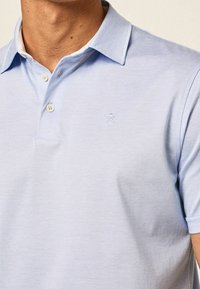 Polo shirt bleu clair avec un col, patte de boutonnage à deux boutons et un petit logo brodé sur le côté gauche. Texture lisse.