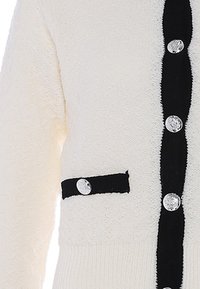 Cárdigan blanco texturizado con una tapeta de botones negra, detalle de bolsillo en contraste y botones plateados redondos. Tejido de punto suave.