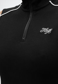 Μαύρο αμάνικο τοπ με φερμουάρ, με λευκή ρέλι στους ώμους και λευκό κεντημένο λογότυπο "Hugo" στο στήθος.