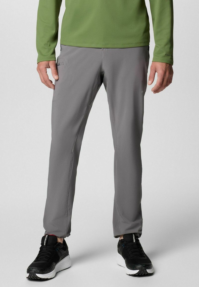 Pantalons de sport gris en tissu léger avec une coupe ajustée. Comprend des poches latérales et des poignets élastiques. Portés avec des baskets noires.