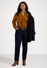 Καφέ μπλούζα με ρυτίδες, σκούρα τζιν, καφέ δερμάτινα τακούνια, καφές ζώνη και ναυτικές σακάκι. Μοντέλο με σγουρά μαλλιά στέκεται μπροστά σε ανοιχτό φόντο.