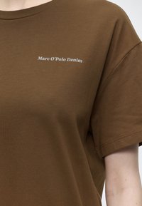 Κοντινό πλάνο ενός καφέ κοντομάνικου T-shirt με λευκή επιγραφή "Marc O’Polo Denim" στο πάνω μέρος του στήθους, που φοριέται από άτομο με ανοιχτόχρωμο δέρμα.