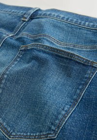 Gros plan sur un jean en denim bleu montrant la poche arrière avec des surpiqûres orange et la boucle de ceinture à la taille.
