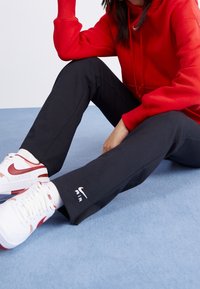 Pantalons de sport noirs à coupe droite, arborant un logo "AIR" blanc sur la jambe. Associés à un hoodie rouge et des sneakers blancs et rouges.